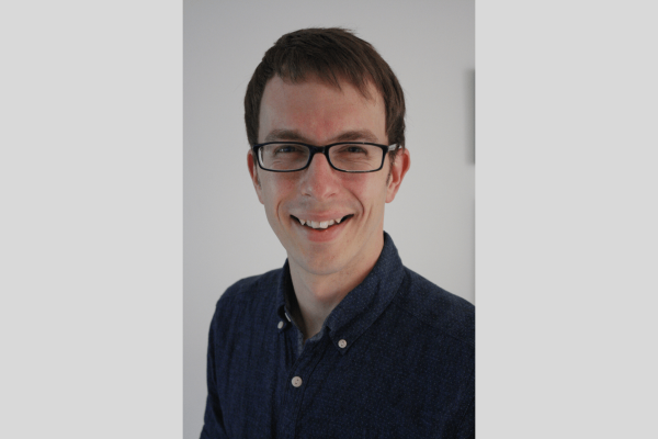 A portrait of Tim Hudson, a white man with short dark hair and glasses, smiling at the camera. He wears a navy shirt. The background is grey